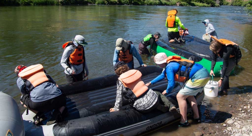 A group of people wearing life jackets appear to clean the bottom of rafts in ankle-deep water.